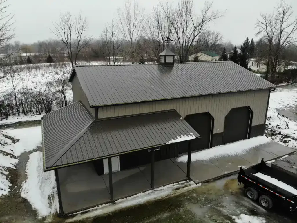 Aerial view of a large, two-toned pole barn with a metal roof and partial snow coverage on the ground, surrounded by leafless trees.