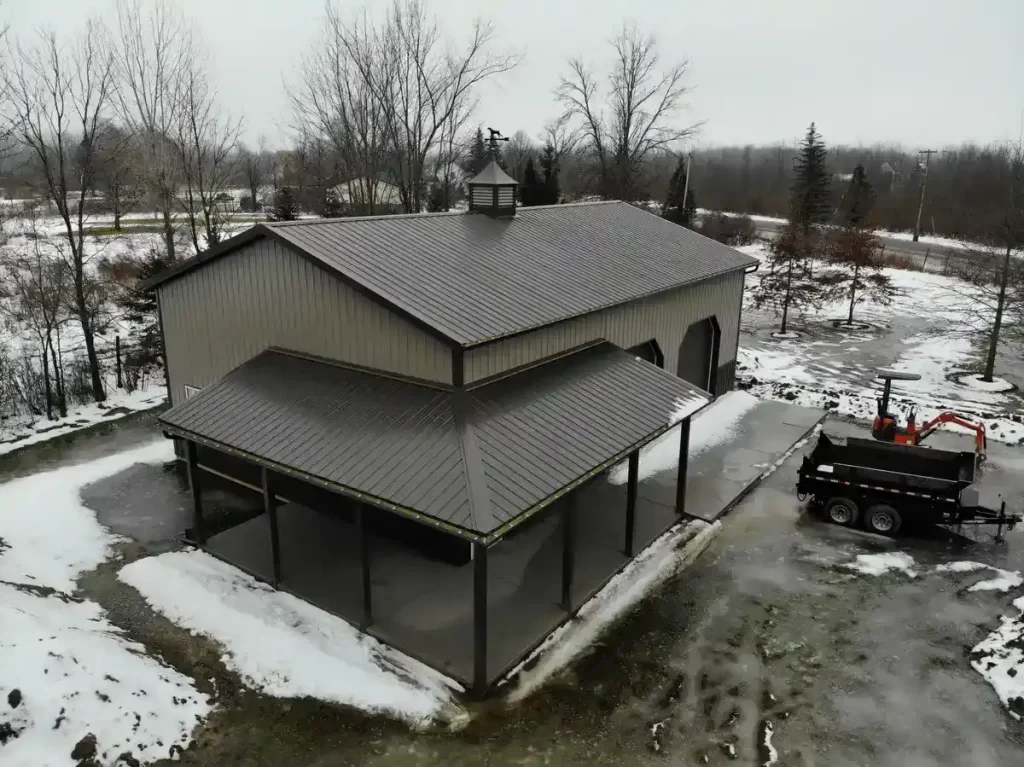 Elevated view of a spacious pole barn with open garage doors and a covered lean-to, with a snowy landscape and construction equipment in the foreground.