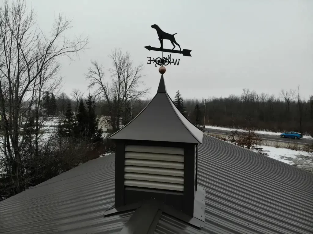 Close-up of a pole barn's cupola and weathervane against a cloudy sky, showcasing detailed metal roofing and vent slats.