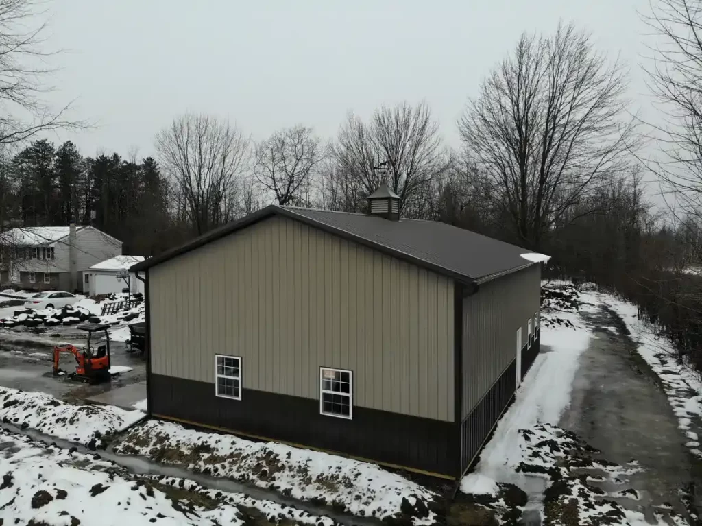 Side angle view of a pole barn in overcast weather, highlighting the contrast between the building's tan walls and the snow-speckled ground