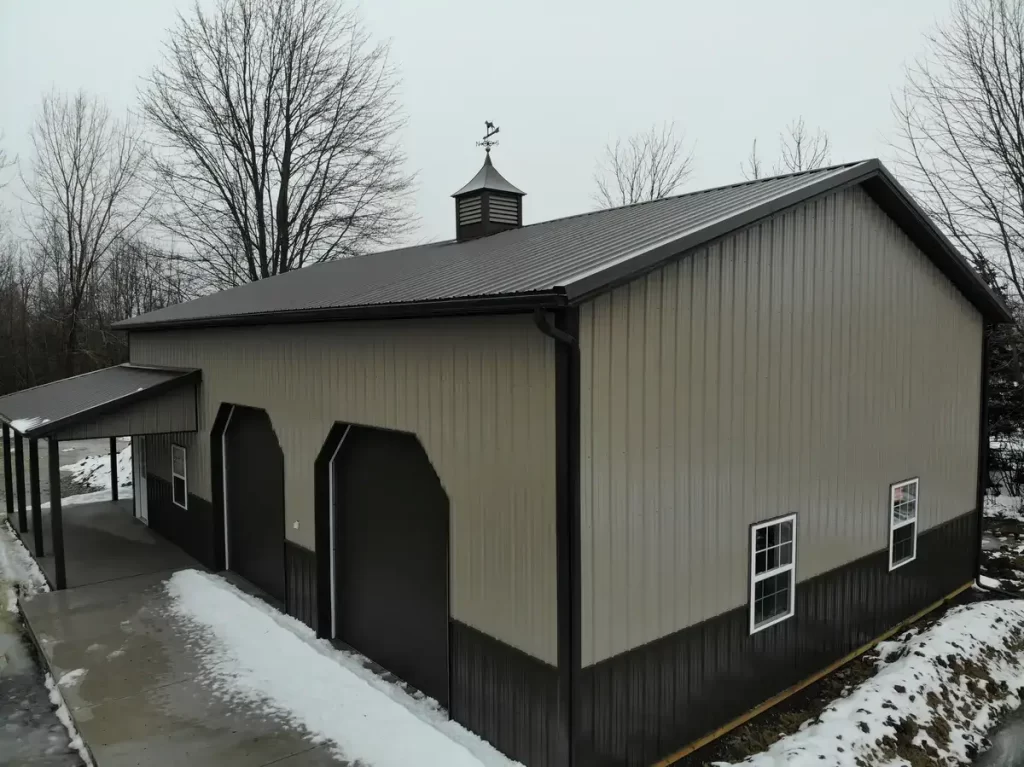 Side view of a pole barn with dark trim, displaying large garage doors and a protective lean-to, on a cloudy day with patches of snow.