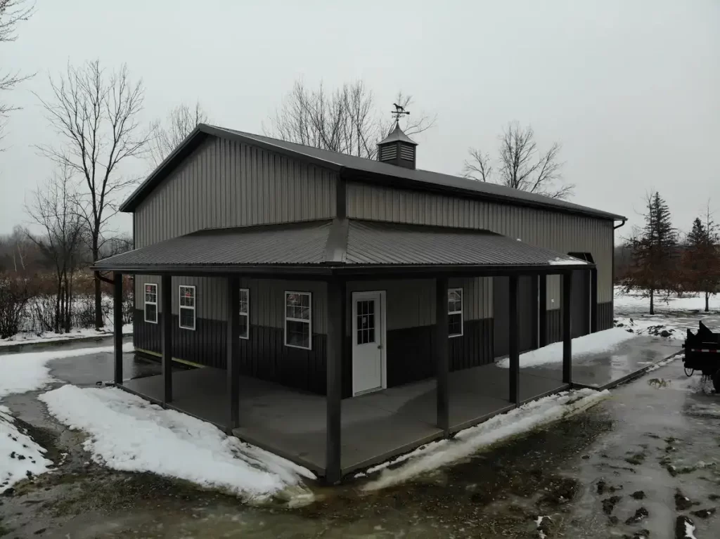 Frontal view of a pole barn with a centered man door, flanked by windows and garage doors, under a grey sky, with a thin layer of snow on the ground.