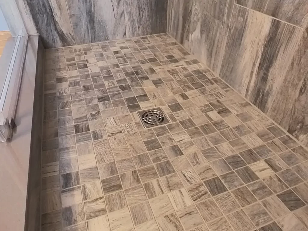 Newly installed shower floor with decorative square mosaic tiles in varying shades of gray and beige, featuring a detailed central drain, part of a high-quality bathroom remodel by Stately Kitchen and Bath, showcasing expert tile work.