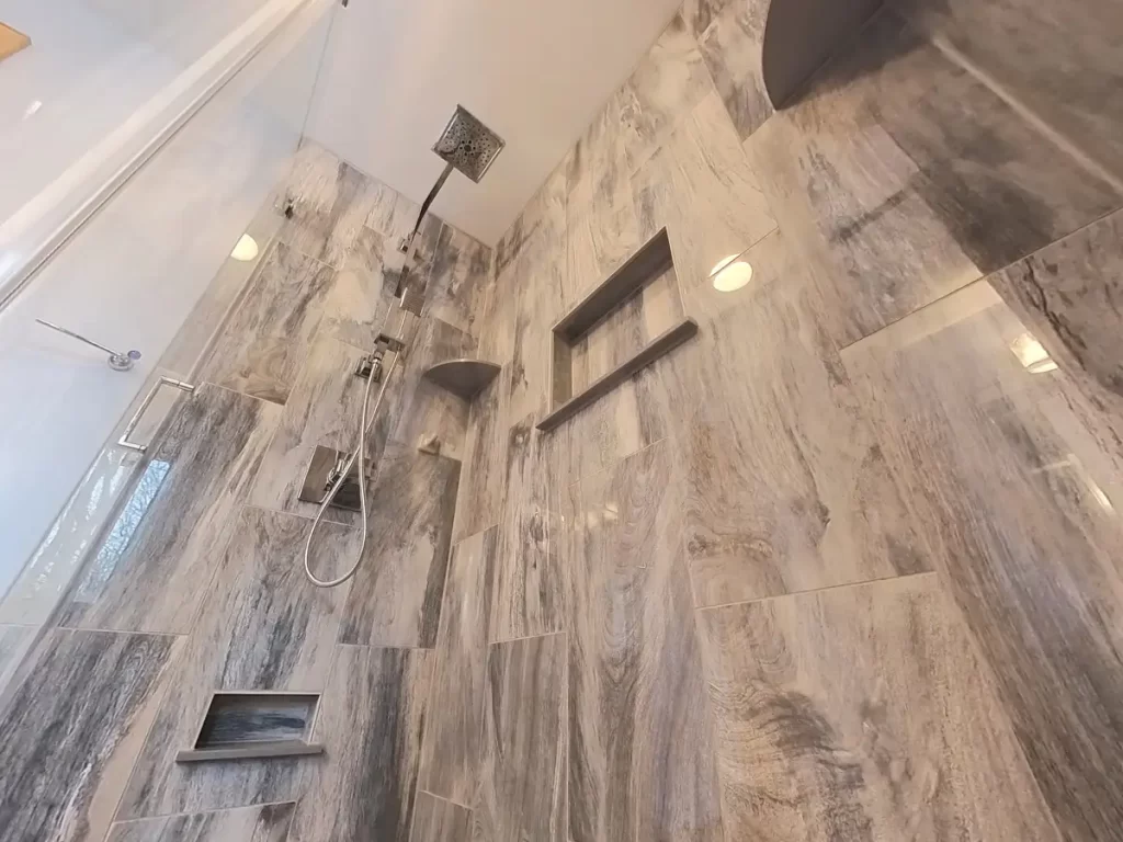 Sophisticated shower interior after a Stately Kitchen and Bath remodel, featuring a wall with gray marble-effect tiles, a sleek handheld shower unit, overhead rain showerhead, and recessed shelves for a clutter-free environment, encapsulating modern design and functionality.