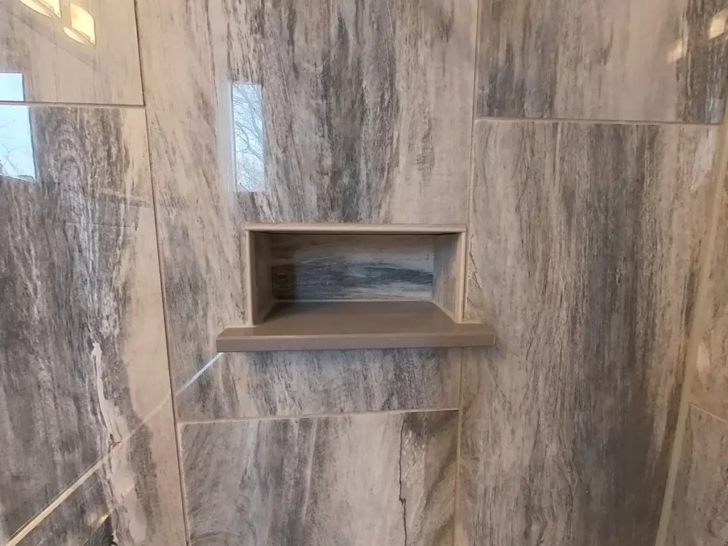 Custom-designed shaving-step shower niche in a marble-patterned tiled wall, a stylish and practical feature of a bathroom remodeled by Stately Kitchen and Bath, showcasing their expertise in adding both beauty and functionality to modern bathroom designs.