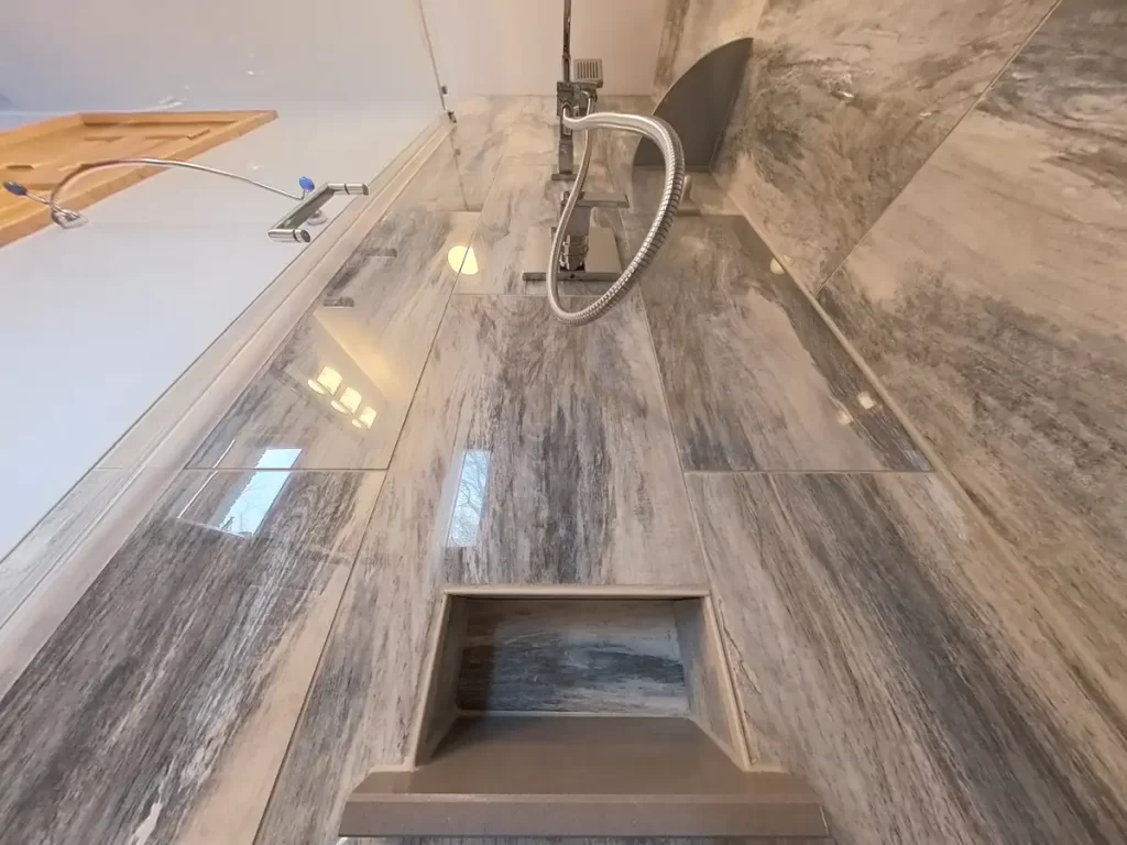Looking up on a luxurious shower space with a contemporary handheld shower fixture, marble-like tiles, and a built-in shaving-shelf wall niche, all part of a high-quality bathroom renovation by Stately Kitchen and Bath, showcasing their commitment to elegant and functional design.