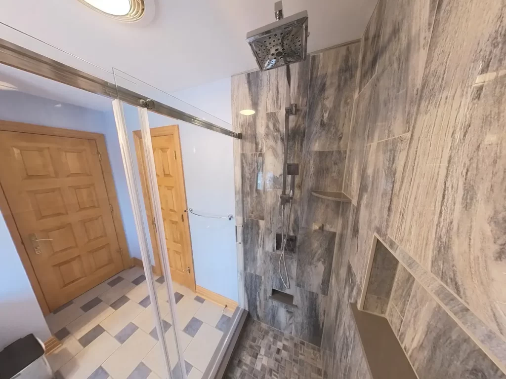 Elegant bathroom after renovation with a clear glass-enclosed walk-in shower, featuring modern gray vein-cut marble tiles, chrome rain showerhead and accessories, and multiple recessed niches for storage, reflecting Stately Kitchen and Bath's upscale remodeling services.