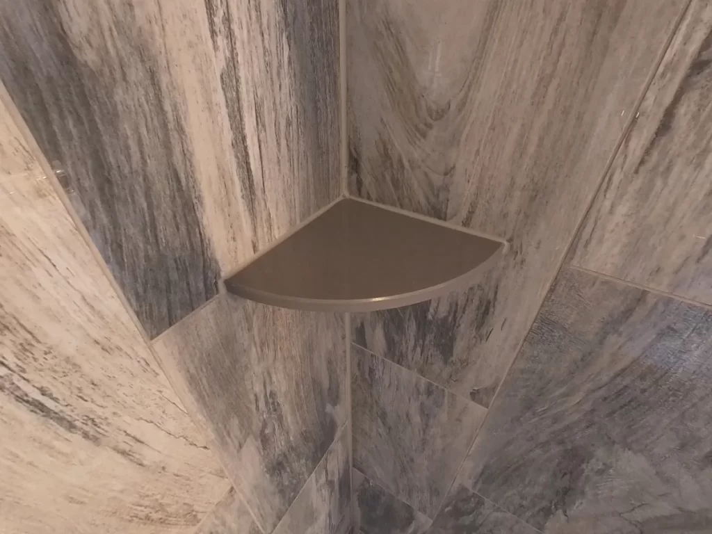 Close-up of a custom-built corner shelf in a newly renovated shower, with gray marble-effect tiles providing a sophisticated backdrop, showcasing Stately Kitchen and Bath's focus on stylish and functional bathroom upgrades.