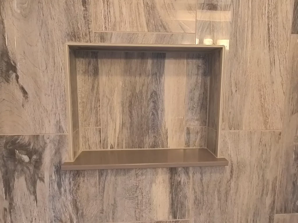 Detail of a modern shower wall niche with a sleek design, framed within gray marble-patterned tiles, illustrating the functional elegance in bathroom storage solutions by Stately Kitchen and Bath.