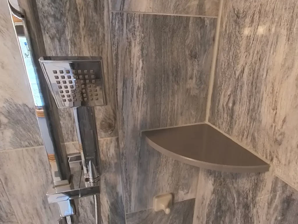 After renovation: A close-up of a contemporary shower fixture with a square handheld showerhead alongside a custom corner shelf, set against the backdrop of elegant gray marble-effect tiles in a remodeled bathroom by Stately Kitchen and Bath.