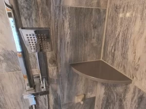 After renovation: A close-up of a contemporary shower fixture with a square handheld showerhead alongside a custom corner shelf, set against the backdrop of elegant gray marble-effect tiles in a remodeled bathroom by Stately Kitchen and Bath.