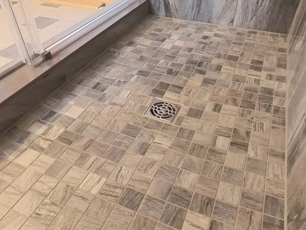 The floor of a newly renovated shower area with a patterned mosaic tile design in shades of gray, featuring a central square drain, demonstrating Stately Kitchen and Bath's attention to detail and craftsmanship in custom bathroom flooring.