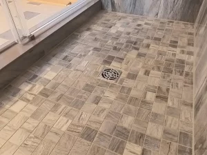The floor of a newly renovated shower area with a patterned mosaic tile design in shades of gray, featuring a central square drain, demonstrating Stately Kitchen and Bath's attention to detail and craftsmanship in custom bathroom flooring.