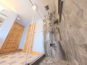 Spacious bathroom after remodel showcasing a luxurious walk-in shower with glass doors, gray marble-look tiles, high-end chrome shower system, and strategically placed niches for toiletries, reflecting Stately Kitchen and Bath's excellence in custom bathroom design.