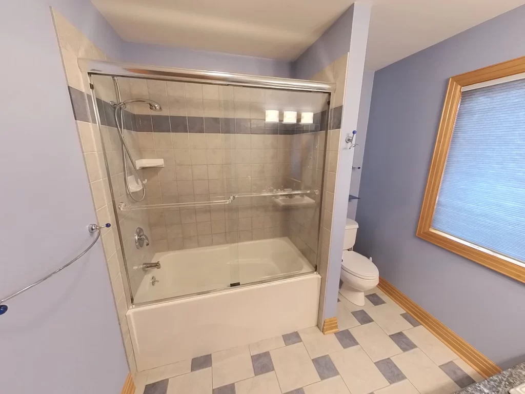 Before renovation: A compact bathroom with a cream-colored bathtub and shower combo, beige wall tiles with a decorative border, silver fixtures, and a single window with a blue shade. The room has a pale blue wall paint and a tiled floor with cream and blue square tiles.