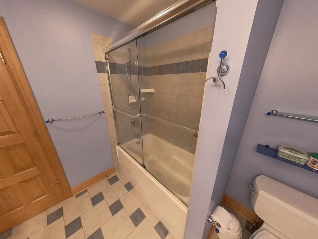 Before renovation: A bathroom corner featuring a bathtub with a clear glass sliding door, cream and beige wall tiles with a blue decorative border, and chrome fixtures. The room has lavender walls, a wooden door, a tiled floor with a diamond pattern in shades of cream and blue, and bathroom safety bars mounted on the wall.
