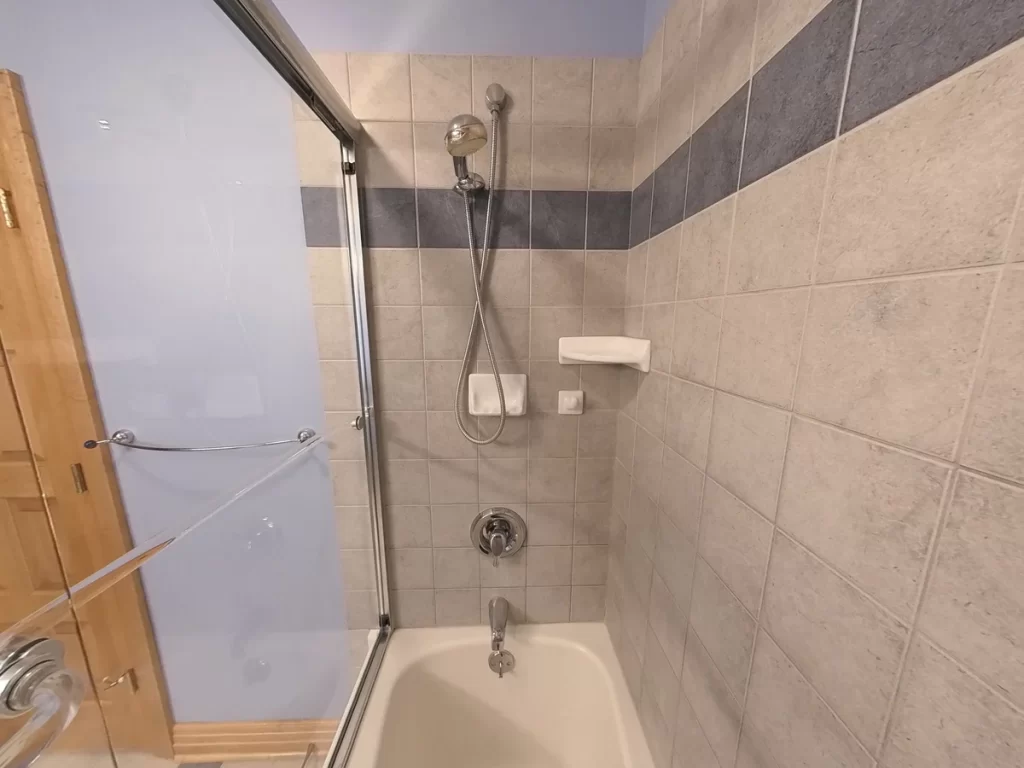 Interior view of a bathroom before renovation, showing a close-up of a bathtub corner with beige wall tiles and a blue accent stripe, a wall-mounted soap dish, and a chrome showerhead and faucet. Visible are the edges of a clear glass door and safety grab bars against a lavender wall.