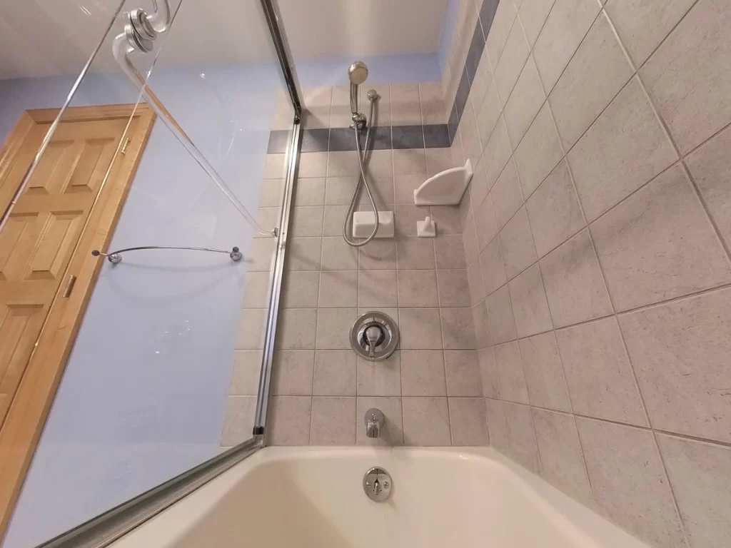 A bathroom before renovation, featuring a cream-colored bathtub with an overhead showerhead, beige wall tiles with a blue decorative tile band, and a glass shower door. To the right is a wooden door, and safety grab bars are installed on the walls.