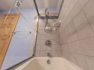 A bathroom before renovation, featuring a cream-colored bathtub with an overhead showerhead, beige wall tiles with a blue decorative tile band, and a glass shower door. To the right is a wooden door, and safety grab bars are installed on the walls.