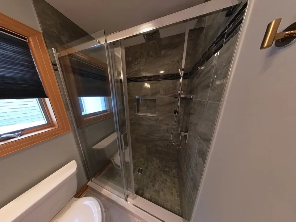Luxurious after-renovation bathroom featuring a glass-enclosed walk-in shower with dark gray tiling, mosaic tile floor, a modern handheld sprayer, and sleek niches for storage, by Stately Kitchen and Bath, against a backdrop of a wooden window frame and neutral walls.