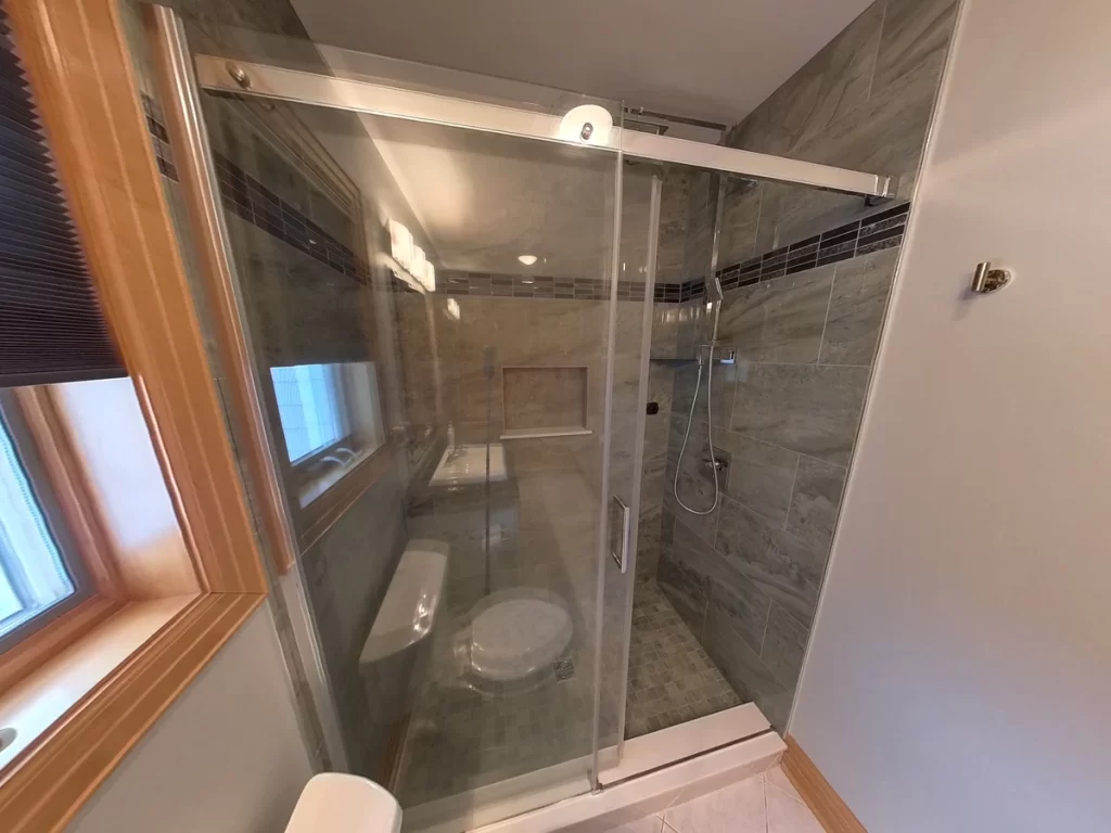 View of a newly remodeled bathroom showcasing a spacious walk-in shower with a clear glass door, dark gray wall tiles with decorative borders, and a contemporary handheld showerhead, part of Stately Kitchen and Bath’s high-quality bathroom upgrades.