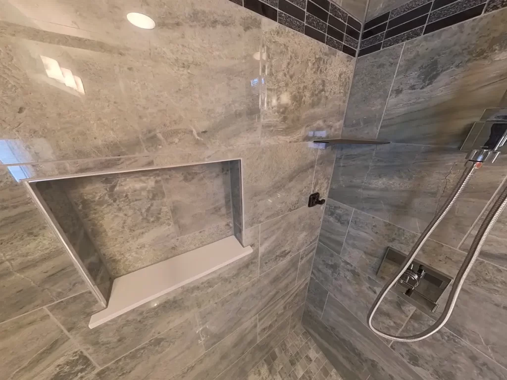 Inside a shower remodeled by Stately Kitchen and Bath featuring gray textured tiles, a custom niche for products, and a chrome handheld shower, all meticulously designed to blend functionality with a sleek, modern aesthetic in a residential bathroom update.