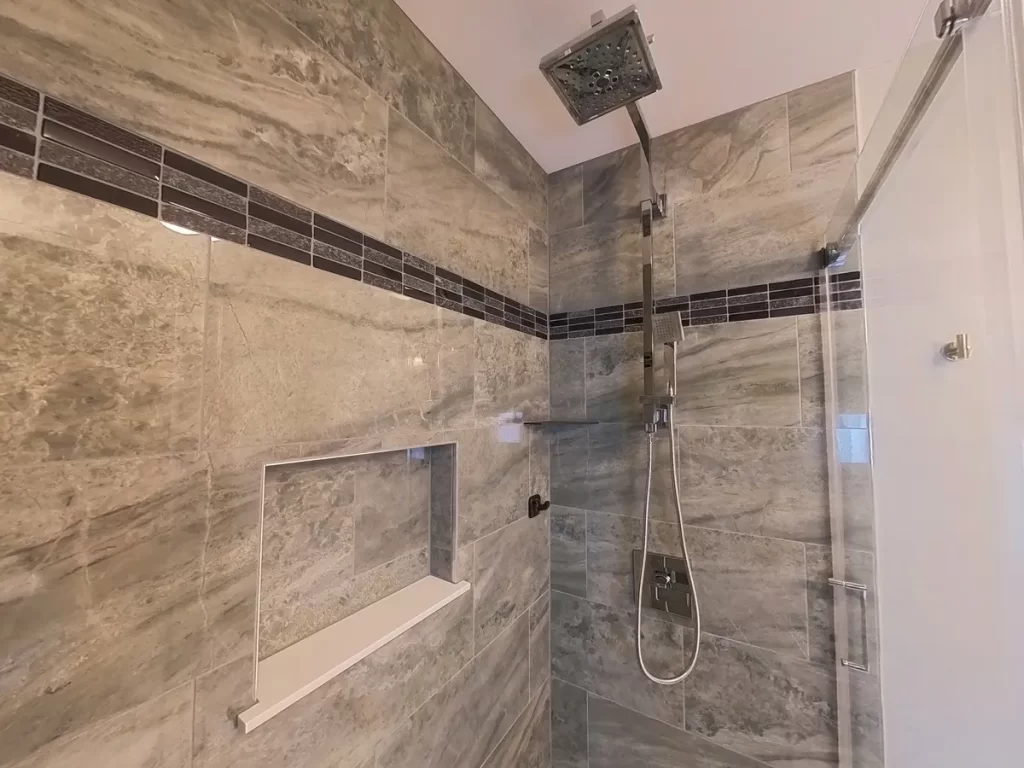 After bathroom remodel, showcasing a walk-in shower with an overhead square rain showerhead, hand-held nozzle, and glass enclosure, with elegant dark gray and decorative border tiles, and a built-in wall niche, displaying the high-quality work of Stately Kitchen and Bath.