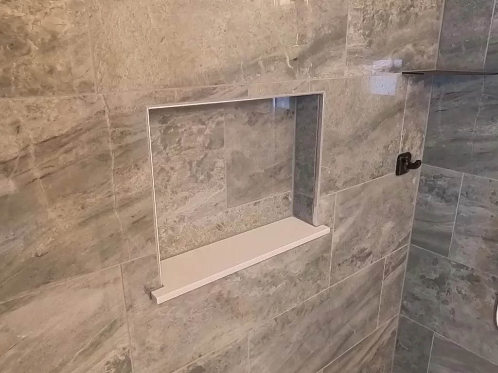 A detailed view of a stylish and functional shower niche in a recently remodeled bathroom, with gray stone-look tiles, providing a sleek and convenient storage solution in a Stately Kitchen and Bath custom-designed modern walk-in shower.