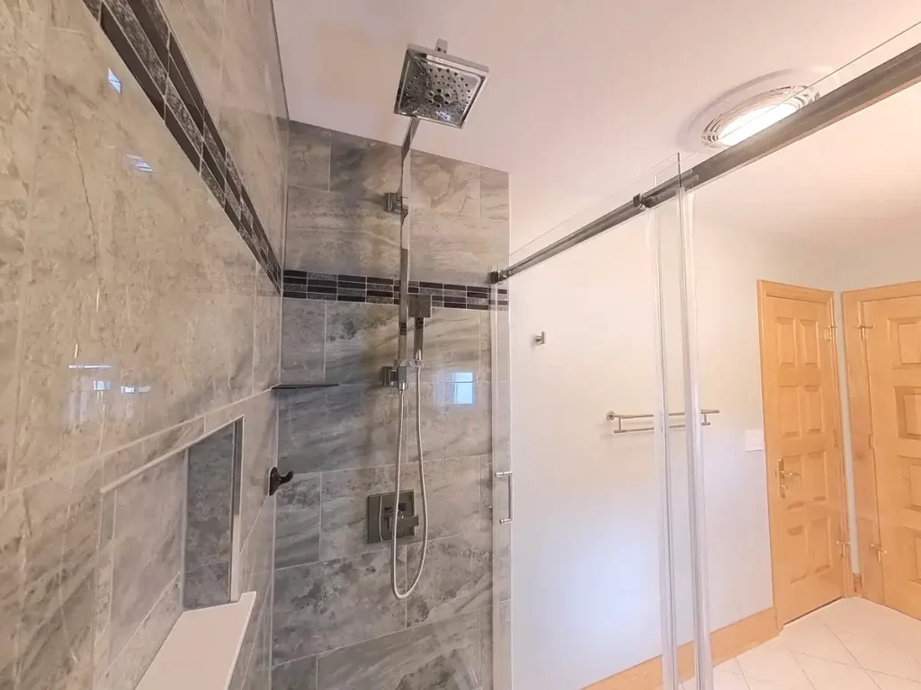 Bright and spacious bathroom showcasing Stately Kitchen and Bath's renovation with a walk-in shower featuring glass doors, a rain showerhead, and gray stone-like tiles, a clear demonstration of modern luxury and functionality in home design.