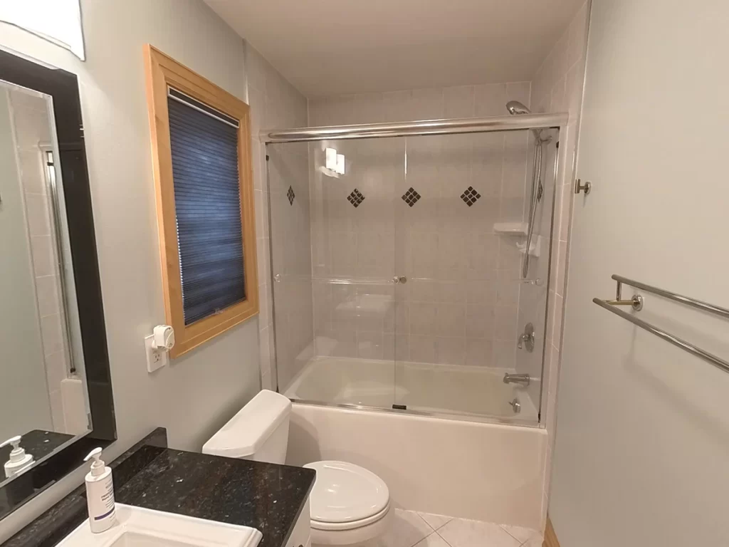 Before image of a bathroom ready for upgrade, featuring a traditional bathtub with a shower and clear glass sliding door, light beige tiles with diamond accents, wooden-framed window with blue blinds, and white walls, exemplifying a typical Stately Kitchen and Bath renovation project.