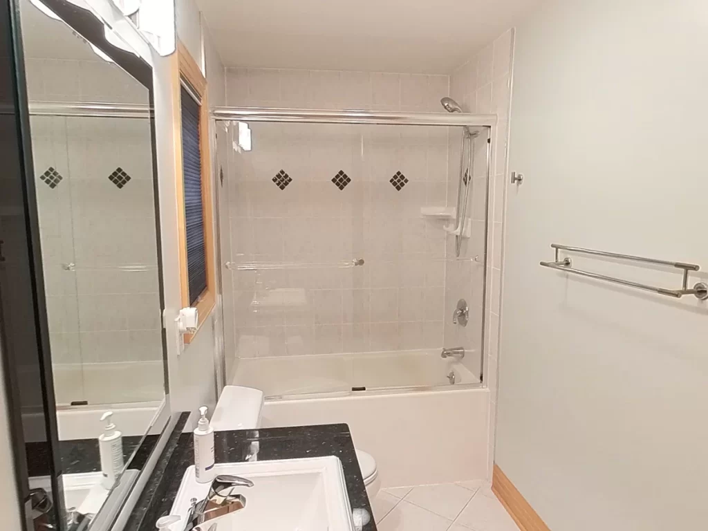 Before renovation bathroom scene with a classic white bathtub and shower combination enclosed by a glass sliding door, neutral-toned wall tiles with decorative diamond-shaped inserts, and essential safety grab bars, all set for a Stately Kitchen and Bath modernization.