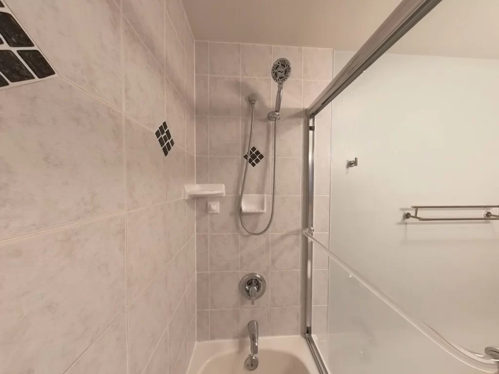 Interior view of a bathroom before remodeling, featuring a classic bathtub with overhead shower, handheld nozzle, and sliding glass doors, surrounded by light tiled walls with black diamond tile accents, ready for a Stately Kitchen and Bath custom upgrade.