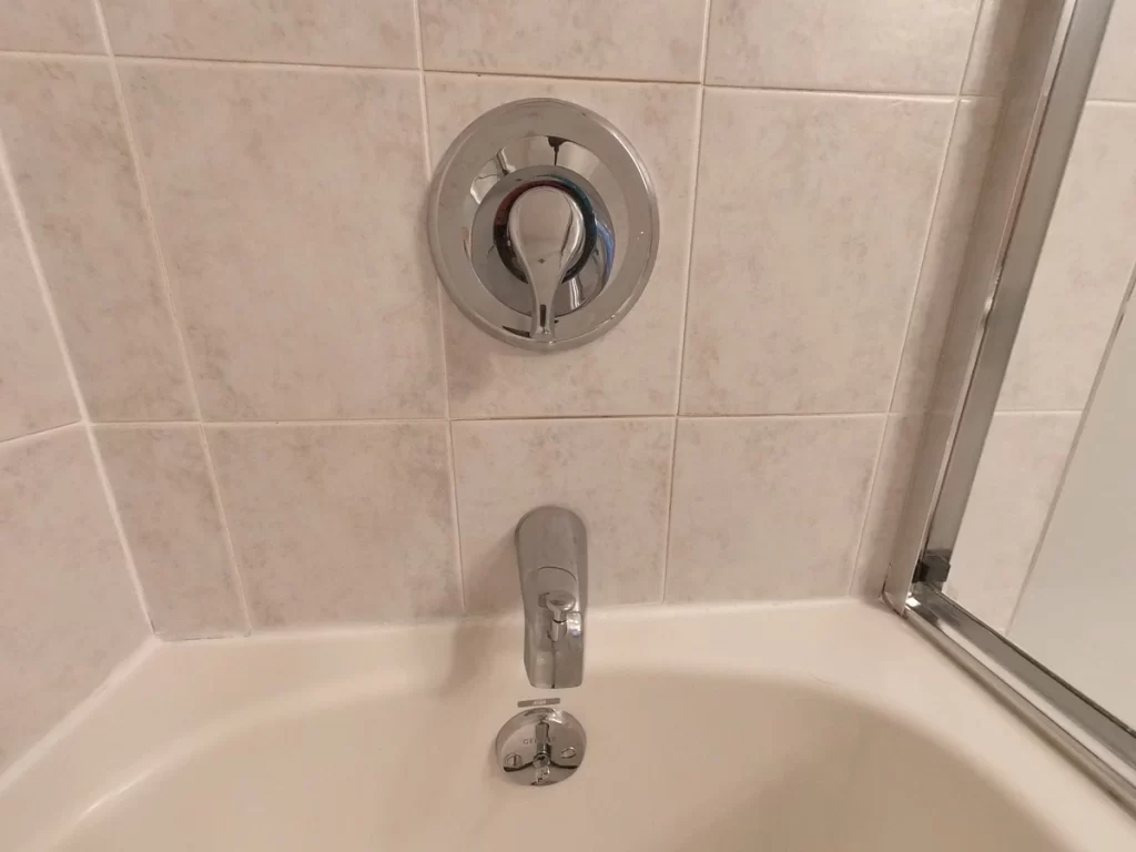 Close-up of bathtub fixtures before renovation, displaying a chrome single-handle faucet and control dial against a backdrop of classic beige wall tiles, representing the starting point for Stately Kitchen and Bath's sophisticated bathroom updates.