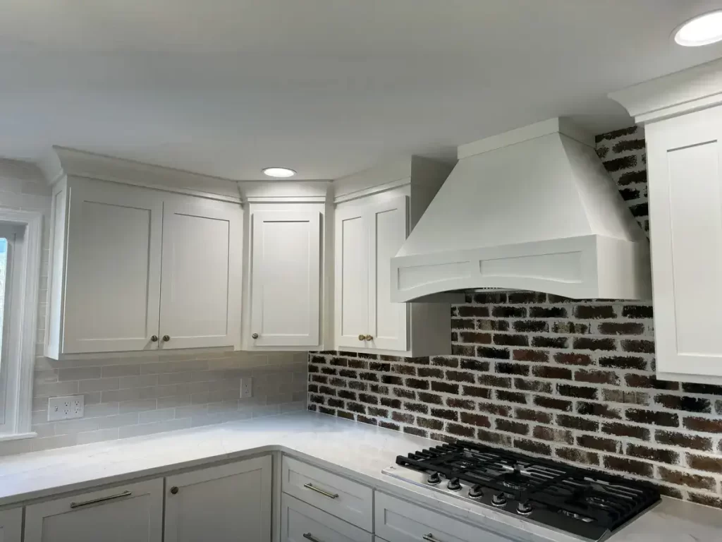Contemporary kitchen remodel in Clarence, NY with exposed brick backsplash, custom white range hood, Reusch Woodworking upper cabinets, and sleek gas stovetop.