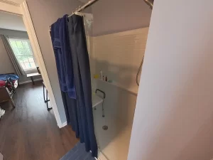 Accessible bathroom in a newly remodeled in-law suite, featuring a walk-in shower with grab bars, blue towels and bath mat, gray curtains, and modern fixtures designed for elder family member care.