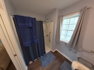 Accessible bathroom in a newly remodeled in-law suite, featuring a walk-in shower with grab bars, blue towels and bath mat, gray curtains, and modern fixtures designed for elder family member care.