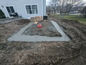 Foundation being laid for a home addition, showing early construction stages with fresh concrete, adjacent to an existing house structure, in a residential backyard setting.