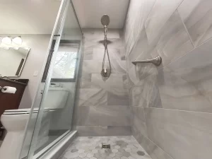 Close-up of a shower with marble tile, rain shower head, and grab bar in a bathroom remodel by Stately Kitchen and Bath in Gettzville, NY, featuring a glass door and hexagonal floor tiles.