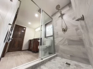 View from inside the shower in a bathroom remodel by Stately Kitchen and Bath in Gettzville, NY, featuring marble tile, a rain shower head, and a glass door.