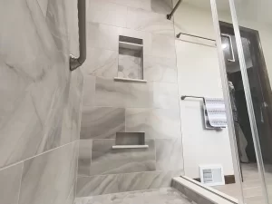 Shower area with built-in shelving and marble tile in a bathroom remodel by Stately Kitchen and Bath in Gettzville, NY, featuring a grab bar and modern fixtures.
