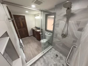 View from inside a remodeled bathroom shower by Stately Kitchen and Bath in Gettzville, NY, featuring a rain shower head, built-in shelving, and a double vanity with granite countertops.