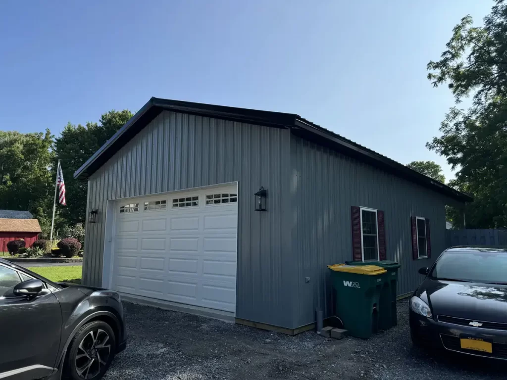 Angled view of a grey post-frame building in Clarence, NY, built by Stately Pole Barns, showing the white garage doors and surrounding property.