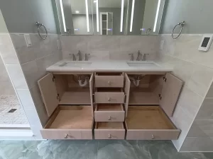 Open vanity drawers in a modern bathroom, showcasing organized storage space beneath dual sinks, with wall-mounted mirrors and bright lighting. Cabinets by Reusch Woodworking and Restoration. Bathroom remodel by Stately Kitchen and Bath.