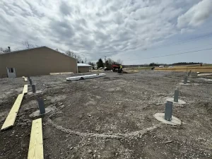Close-up of Perma-Column brackets securely installed into the poured concrete piers at the Bennington Highway Department pole barn construction site, with Stately Builders equipment visible in the background.