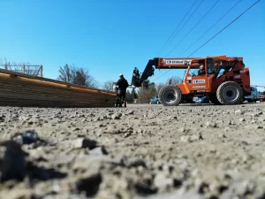 Overhead view of a construction worker and equipment lifting a large truss during the framing phase of the Bennington Highway Department pole barn build, with materials and vehicles in the background.