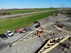 Ground-level view of a construction worker guiding wooden trusses during the framing process of the Bennington Highway Department pole barn, with a forklift in operation beside the stacked materials.