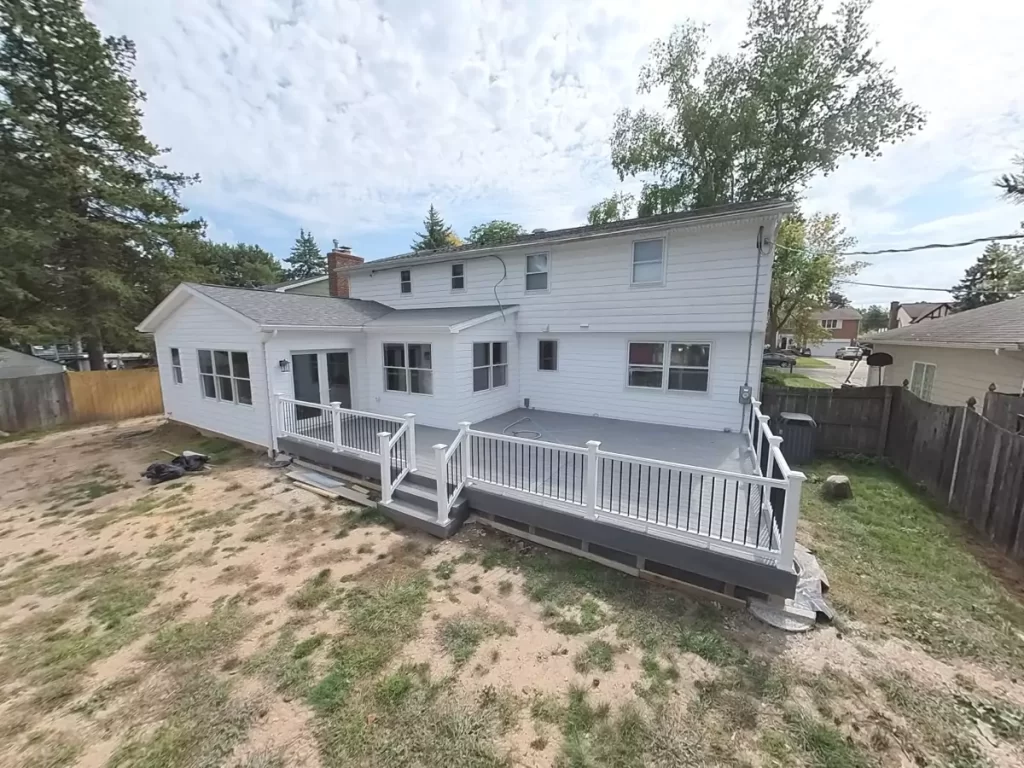 Angled view of a renovated white home with a stylish deck and black railing, highlighting an expanded outdoor space for gatherings and relaxation.