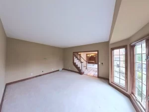 Amherst NY living room pre-remodel with bay window, beige carpet, neutral walls, and open view to staircase and dining area.
