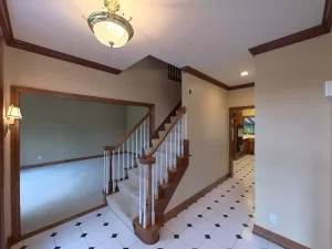 Amherst NY foyer pre-remodel with tile flooring, wood-trimmed staircase, crown molding, and view into kitchen.