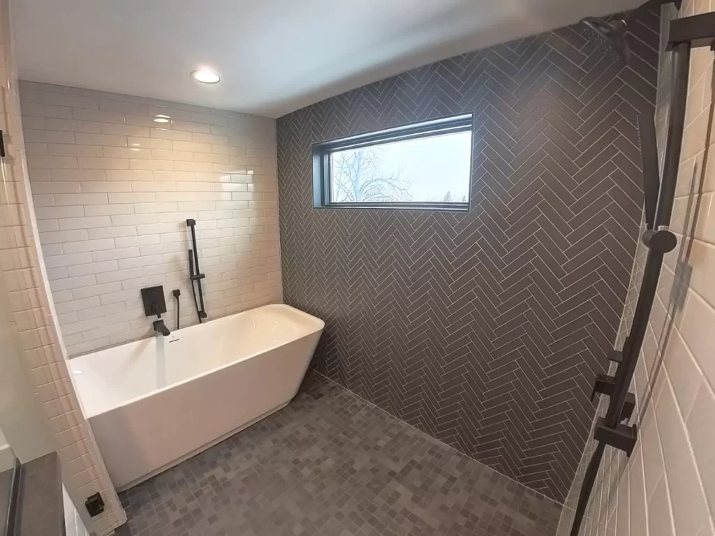 Freestanding tub with black fixtures and full herringbone tile accent wall beneath transom window in Buffalo custom bathroom.